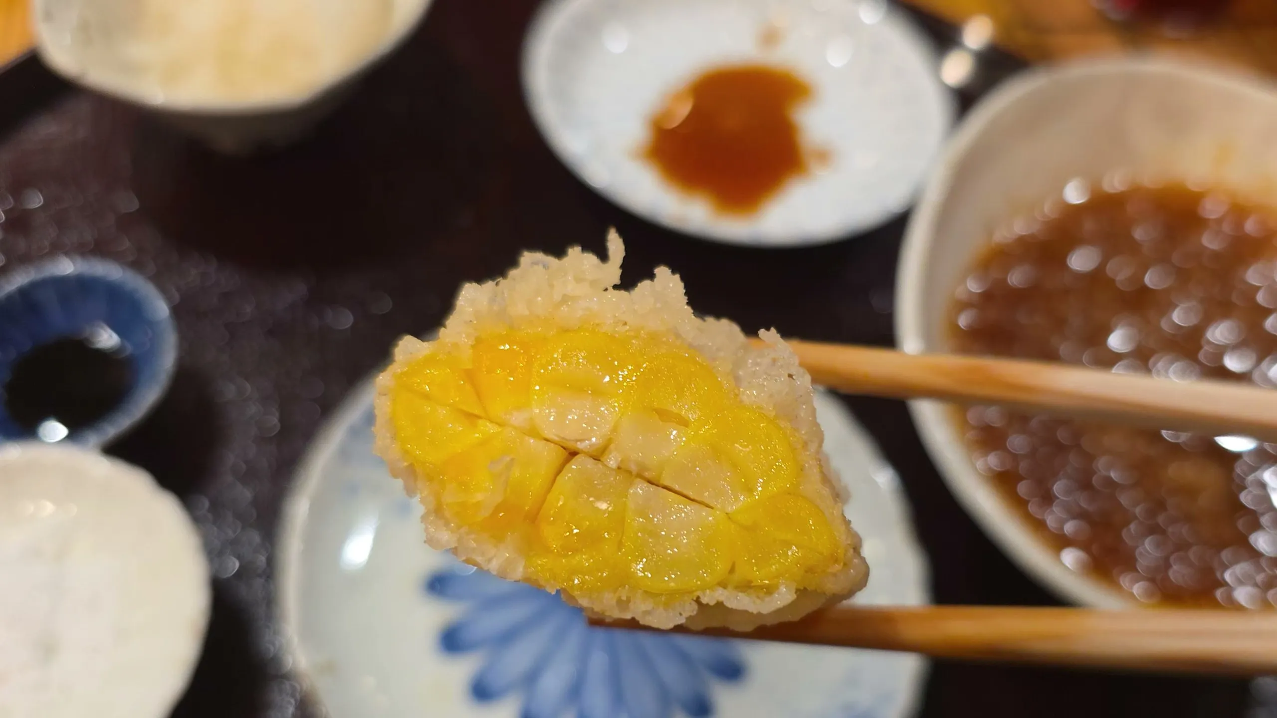 A piece of corn tempura held in chopsticks over dipping sauces