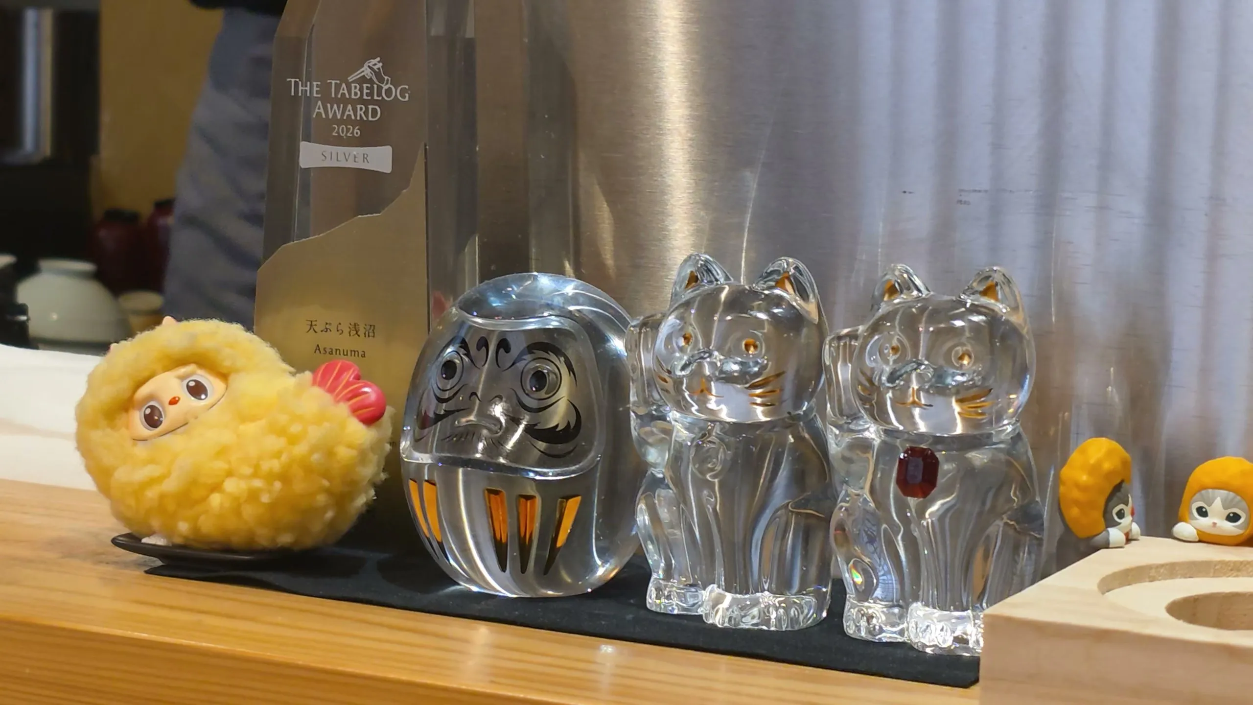 A shrimp-tempura Labubu figure, glass daruma and cats, and a Tabelog Award 2026 Silver trophy on the Asanuma counter